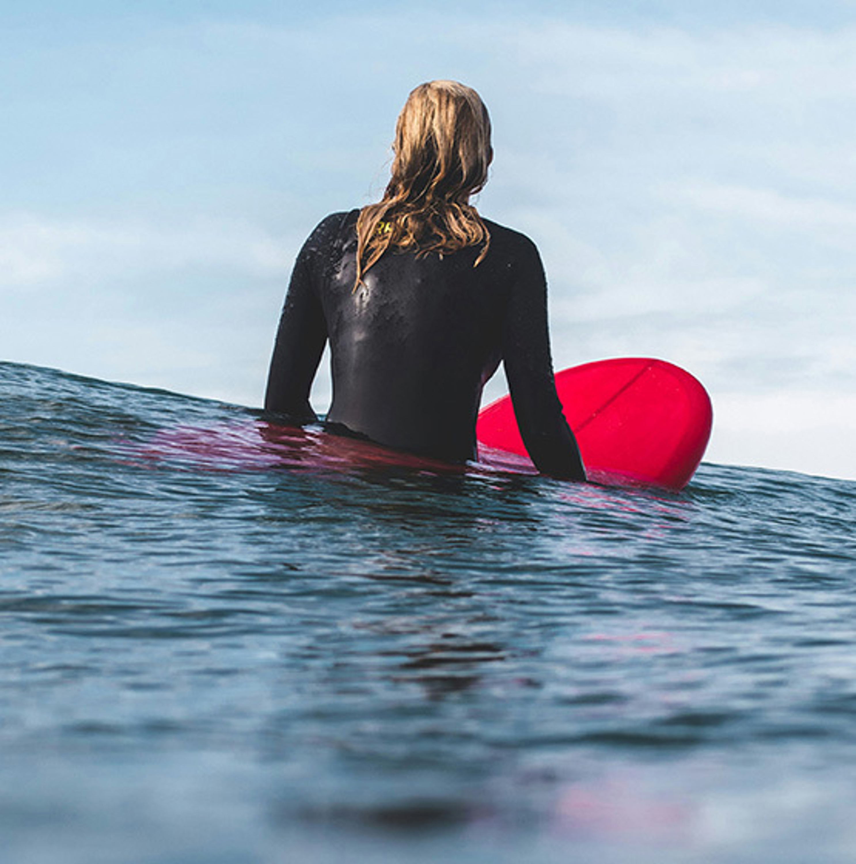 surfer sitting on a red surfboard in the water wearing a long sleeve wetsuit